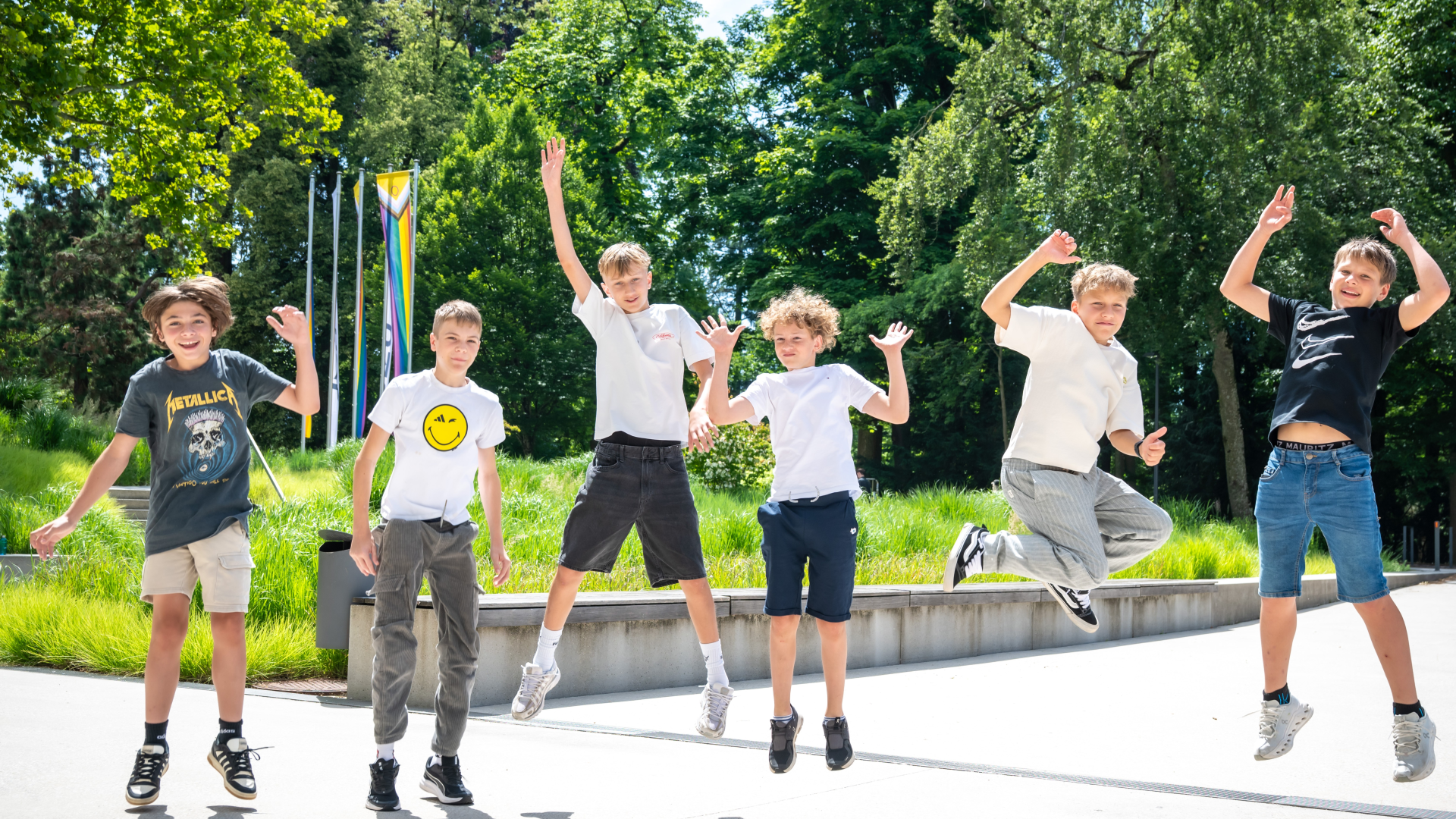 Sechs Schüler:innen springen fröhlich in die Luft vor grüner Wiese, Regenbogenfahne im Hintergrund